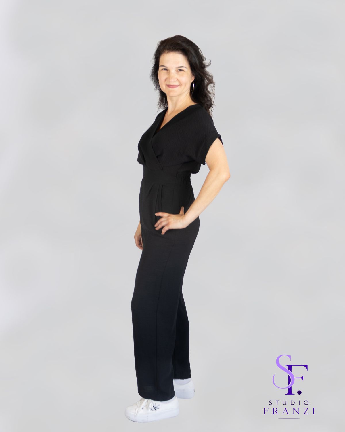 Authentisches Ganzkörperportrait einer Frau im schwarzen Outfit mit selbstbewusster Pose vor neutralem Studiohintergrund.