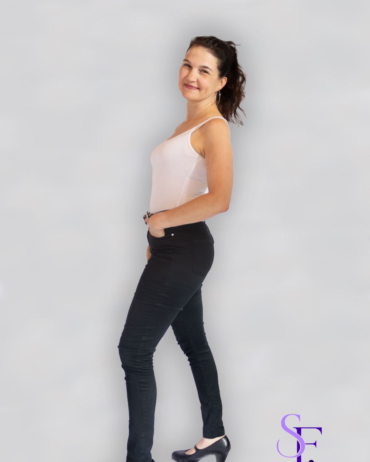 Seitliches Ganzkörperportrait einer lächelnden Frau in weißem Top und schwarzer Hose vor hellem Studiohintergrund.