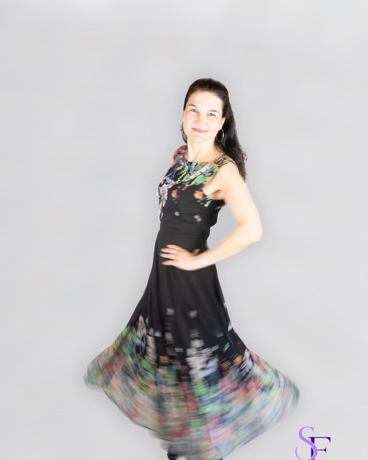 Portrait einer lächelnden Frau in einem schwarzen Kleid mit Blumenmuster während einer Drehbewegung vor hellem Studiohintergrund.