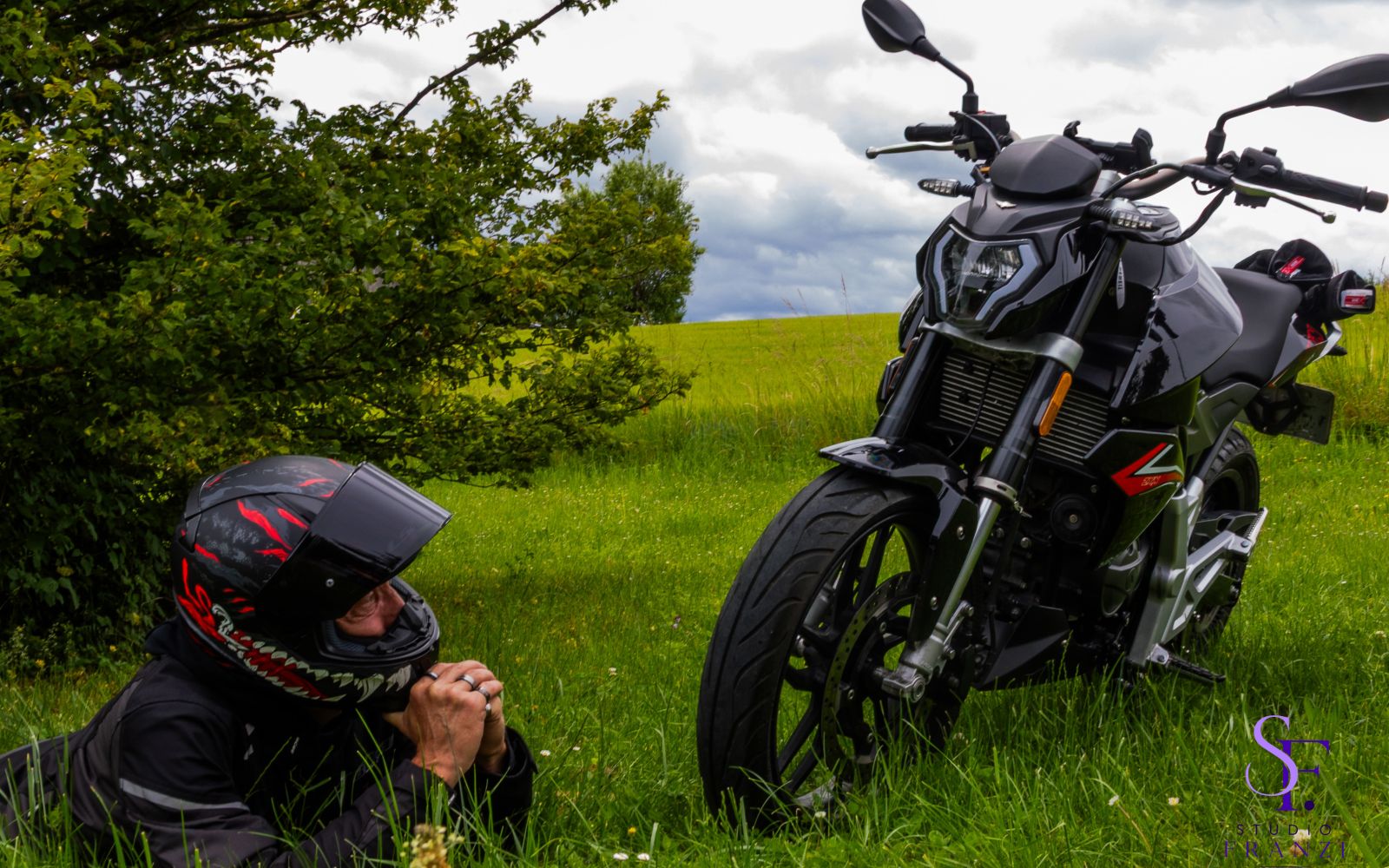 Motorradfahrer liegt im Gras neben einem schwarzen Motorrad auf einer grünen Wiese unter bewölktem Himmel.