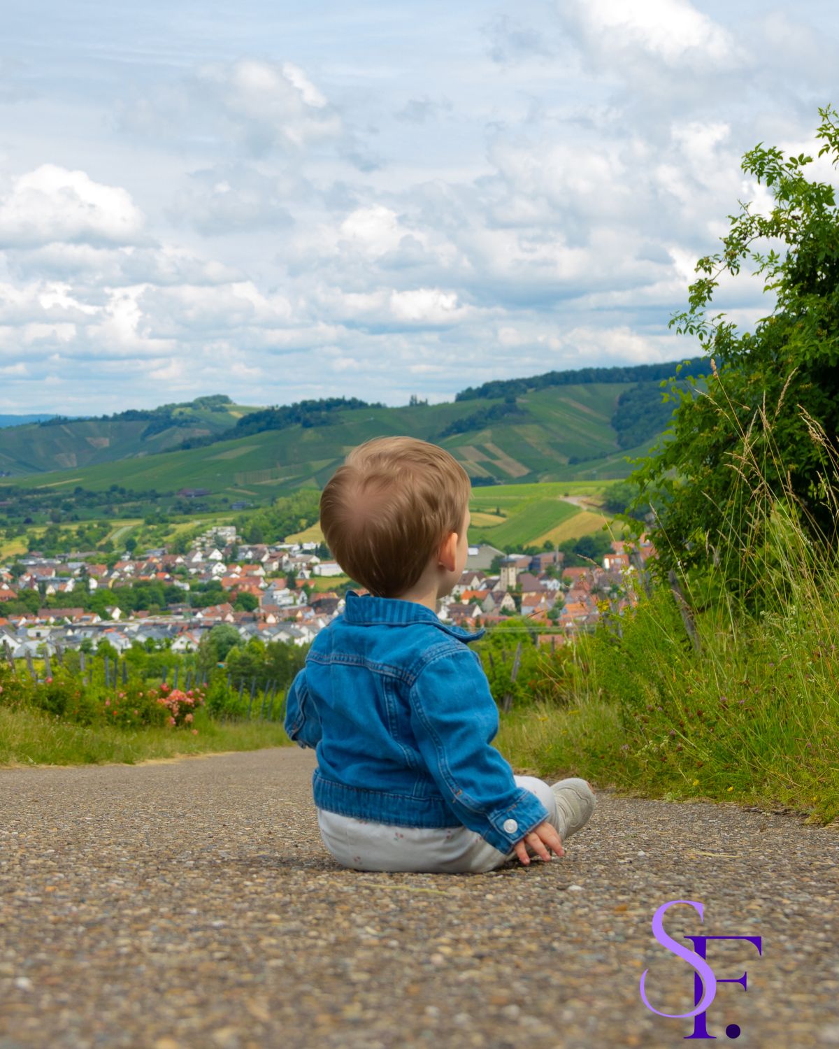 Baby sitzt auf einem Weg und blickt auf eine Weinlandschaft mit Dorf und bewölktem Himmel.