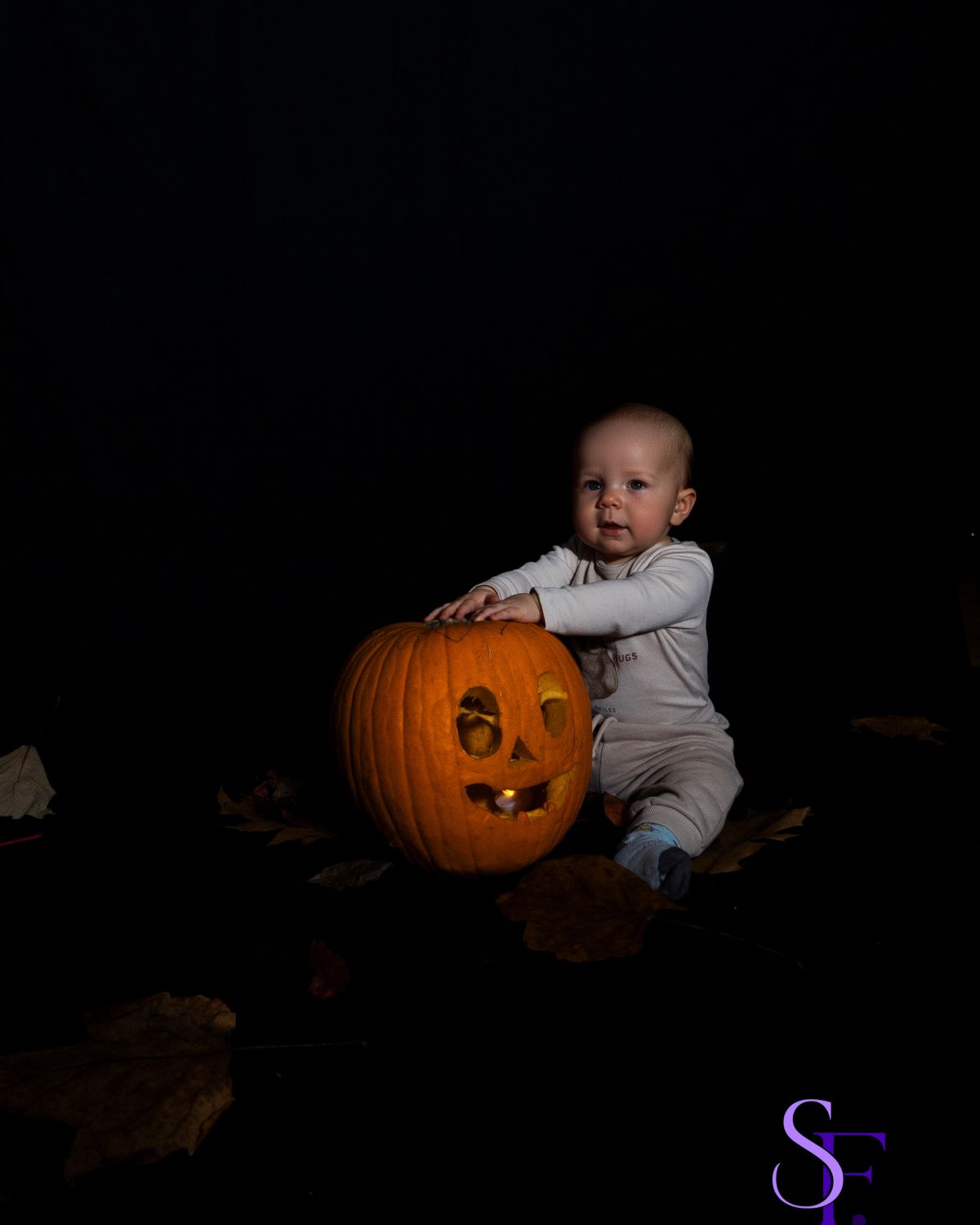 Baby sitzt neben einem leuchtenden Halloween-Kürbis vor schwarzem Studiohintergrund.